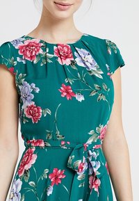 Květinové šaty ze zelené látky s krátkými rukávy, skládacími výstřihy a páskem v pase. Mezi detaily jsou růžové, červené a modré květiny.