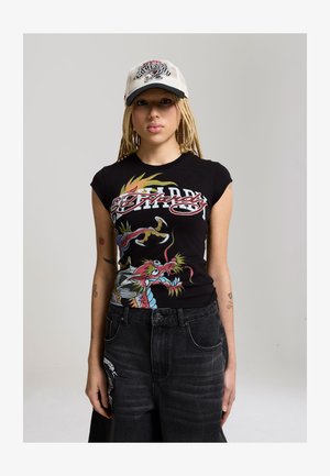 Jonge persoon met gevlochten haar, een witte pet en een zwart grafisch T-shirt met een kleurrijk draakontwerp en een donkere spijkerbroek.