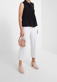 Mujer vestida con una blusa negra sin mangas, pantalones cortos blancos, mule con tacón nude y sosteniendo un pequeño bolso rosa a su lado.
