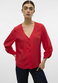 Blusa rossa in tessuto leggero con scollatura a V, chiusura a bottoni frontale e dot texture. Maniche lunghe arrotolate ai polsini.