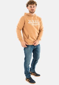 Sudadera color marrón con logo blanco, bolsillo frontal, combinada con jeans azules y zapatillas navy con detalles en marrón, de pie sobre un fondo blanco.