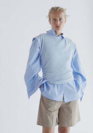 Mujer con camisa azul clara de capas con mangas extragrandes y pantalones cortos beige hasta la rodilla, de pie frente a un fondo liso.