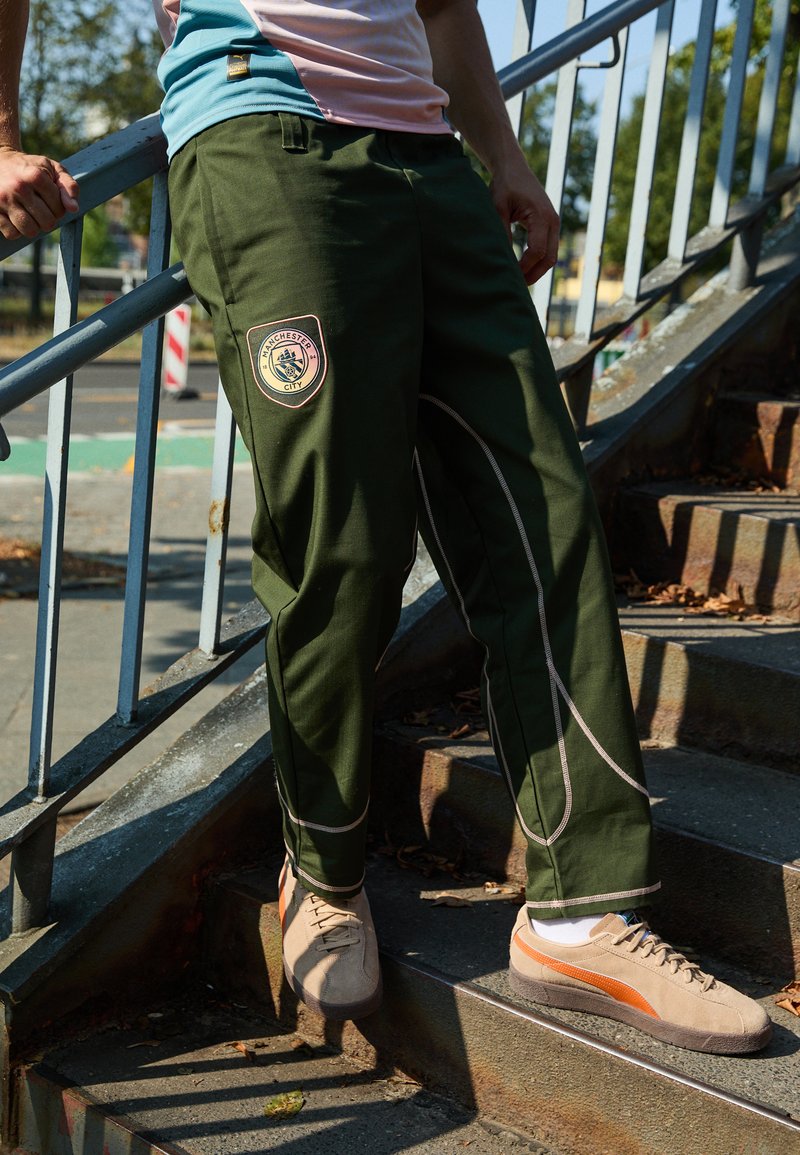 Grüne Sporthosen mit einem Manchester City-Patch, versehen mit weißen Nähdetails und einem lockeren Schnitt. Kombiniert mit beigen und orangefarbenen Sneakers.