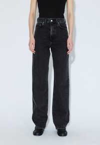 Persona che indossa jeans neri a vita alta larghi, un top nero e scarpe nere, in piedi contro uno sfondo chiaro e uniforme.