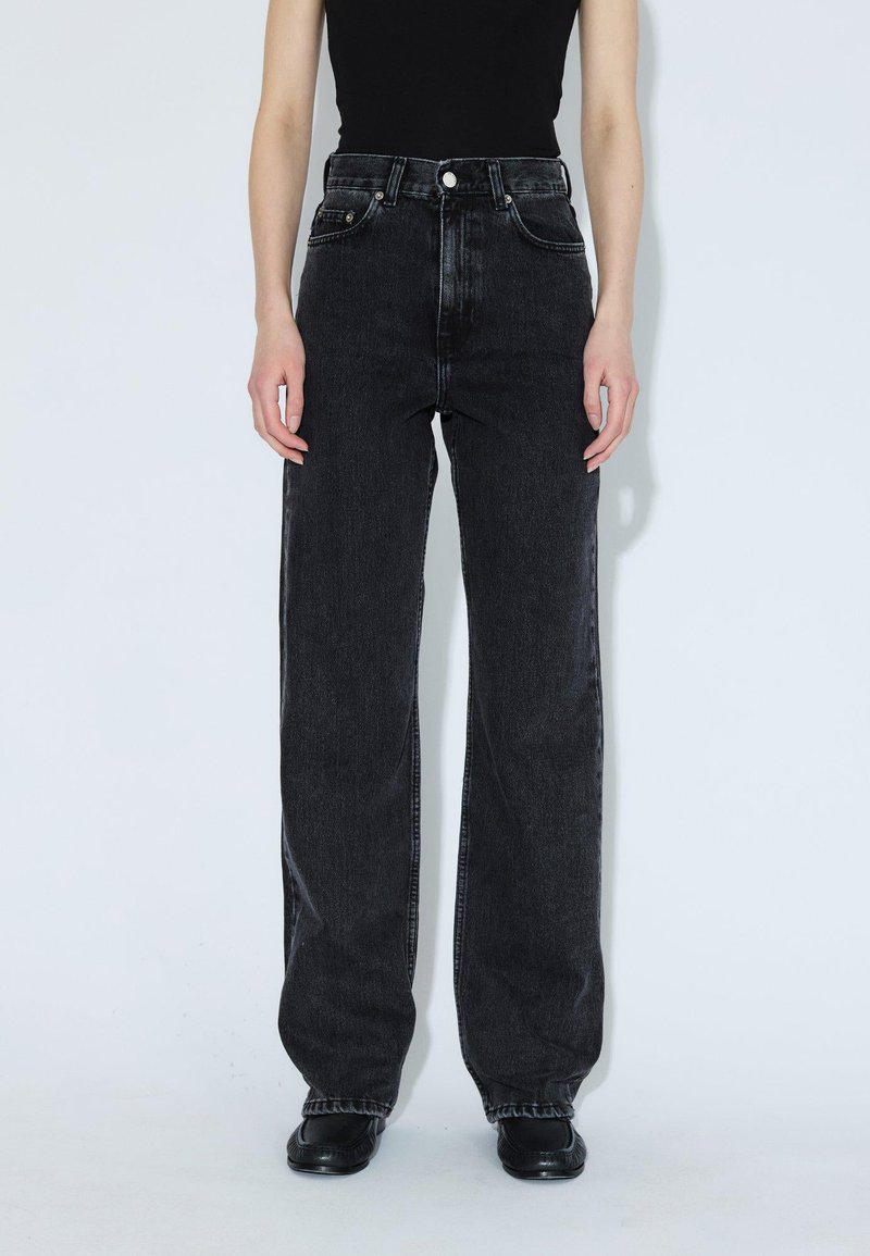 Persona che indossa jeans neri a vita alta larghi, un top nero e scarpe nere, in piedi contro uno sfondo chiaro e uniforme.