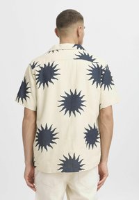 Camicia a maniche corte con bottoni in tessuto beige, caratterizzata da grandi motivi a raggi di sole neri, con colletto aperto e orlo dritto.