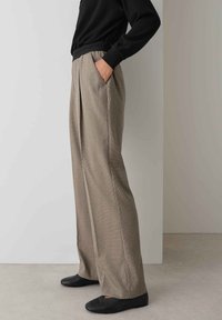 Weite Hosen aus einem strukturierten braunen Stoff, mit vorderen Falten und seitlichen Taschen. Kombiniert mit einem schwarzen Oberteil und flachen schwarzen Schuhen.