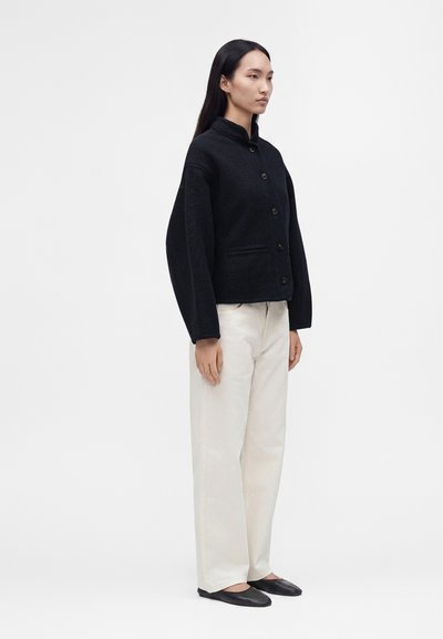 Femme debout portant une veste noire boutonnée, un pantalon blanc à jambes larges et des ballerines noires, regardant vers la droite sur un fond clair uni.