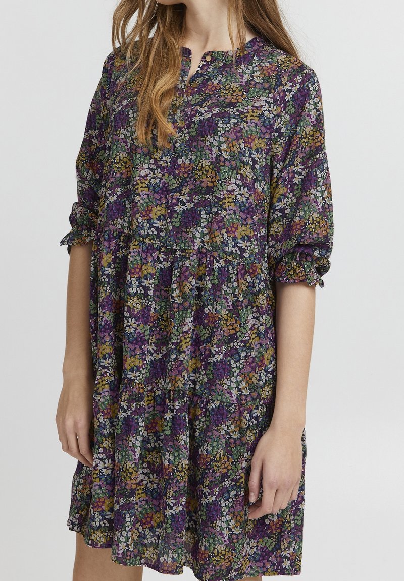 Femme portant une robe fleurie ample avec des fleurs violettes, vertes, jaunes et blanches, avec des manches trois-quarts munies de poignets froncés.