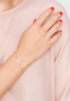 Rose gold bracelet featuring a wavy design with small gems, attached to a delicate chain, worn on a hand against a soft pink fabric background.