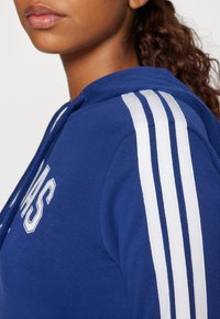 Mladý člověk v modré mikině s bílými pruhy na rameni a částečným bílým nápisem na hrudi, s vlasy svázanými zpět.