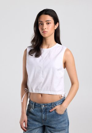 Jonge vrouw met lang donker haar, die een witte mouwloze cropped top en een blauwe spijkerbroek draagt, staand tegen een effen grijze achtergrond.