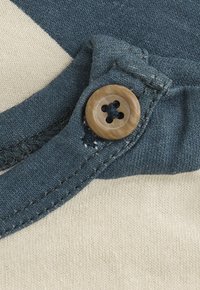 Nahaufnahme eines Holzknopfes, der an einem blauen Stoffriemen über einem cremefarbenen Strickstoff befestigt ist und die Stichdetails zeigt.