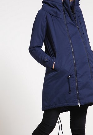 Femme portant une longue veste à capuche bleu marine avec fermeture éclair à l'avant et poches, associée à un pantalon noir.