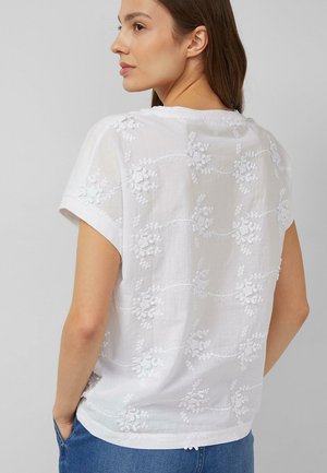 Woman with brown hair wearing a white short-sleeve top with floral embroidery, standing with hands in blue jeans pockets, facing sideways.