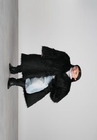 Γυναίκα φορώντας ένα μαύρο μακρύ γούνινο παλτό, ανοιχτό μπλε τζιν, μαύρες μπότες και ένα ανοιχτόχρωμο πουλόβερ με λαιμόκοψη, στέκεται μπροστά σε ουδέτερο φόντο.