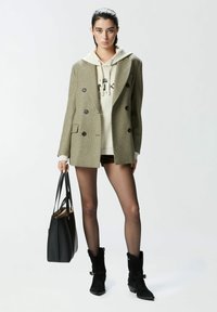 Blazer doppiopetto verde oliva sopra un hoodie beige, abbinato a stivali neri con tacco e una borsa nera a tracolla. Tessuto liscio, vestibilità su misura.