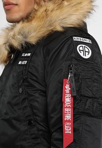 Veste bomber noire avec capuche en fausse fourrure, détails en patch sur la manche et étiquette rouge "REMOVE BEFORE FLIGHT".