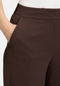 Pantalon large marron en tissu lisse. Les caractéristiques comprennent une taille plate et une poche latérale, avec des coutures nettes et un design soigné.