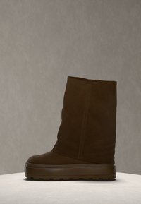 Botte en daim marron mi-mollet avec une épaisse semelle en caoutchouc et un bout arrondi, posée sur une surface claire devant un fond beige uni.