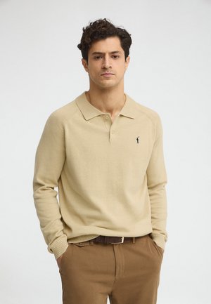 Homme aux cheveux bouclés et foncés, portant un pull-polo beige à manches longues avec un petit logo bleu marine et un pantalon marron avec une ceinture, mains dans les poches.