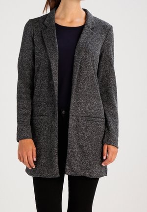 Femme portant un blazer texturé gris foncé sur un haut noir et un pantalon noir, debout devant un fond clair uni.