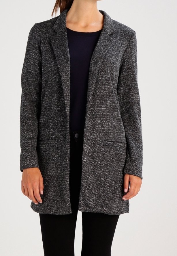 Femme portant un blazer texturé gris foncé sur un haut noir et un pantalon noir, debout devant un fond clair uni.