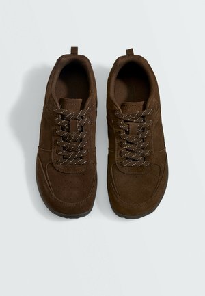 Pair of brown suede sneakers with patterned laces, perforated toe area, and pull tabs at the back on a plain light background.