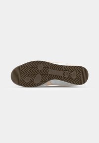 Semelle d’une basket avec une bande de roulement en caoutchouc marron présentant des motifs en zigzag et circulaires sur un fond blanc uni.