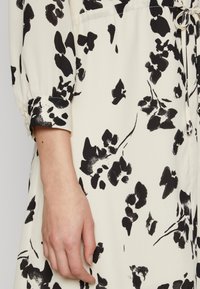 Mustavalkoinen kukkakuosisempi paitapusero pehmeästä kankaasta, jossa on nyöri vyötäröllä ja napitettavat hihat.