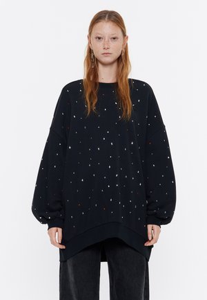 Sudadera negra de gran tamaño con acentos de pedrería dispersos, puños acanalados y un dobladillo curvado. Combinada con pantalones oscuros.