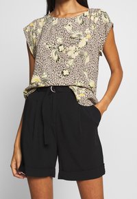 Blus med blommönster i beige, gult och grönt, med korta ärmar. Matchad med högmidjade svarta shorts med knytskärp.