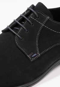 Chaussure en suede noire à lacets avec des coutures blanches contrastantes, bout rond et semelle en caoutchouc lisse. Elle possède quatre œillets et une finition texturée.