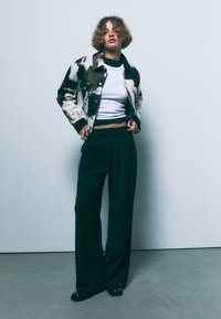 Retro plašč s kravjim vzorcem, narejen iz volne, v beli in rjavi barvi, v kombinaciji z belo majico in širokimi črnimi hlačami. Gladka tekstura, sproščen kroj.