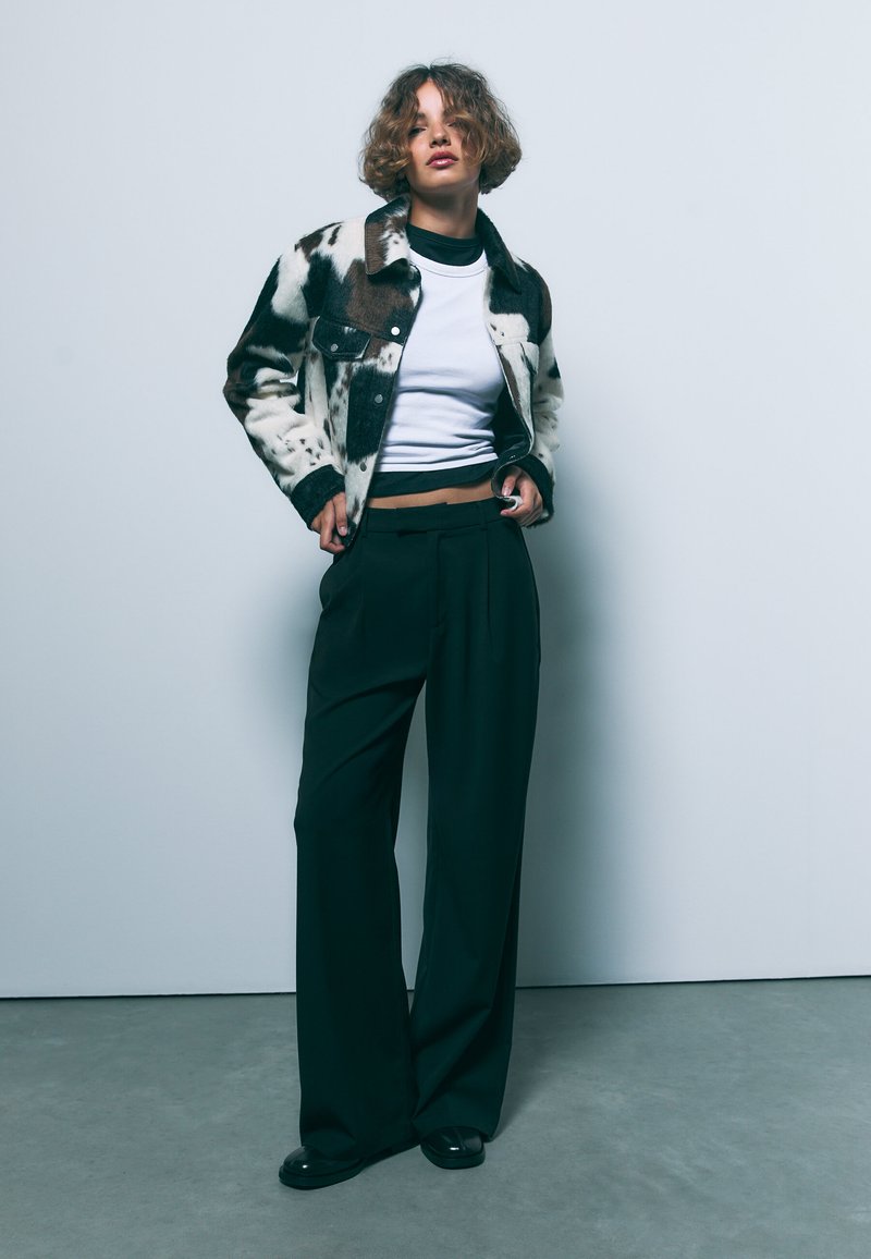 Retro plašč s kravjim vzorcem, narejen iz volne, v beli in rjavi barvi, v kombinaciji z belo majico in širokimi črnimi hlačami. Gladka tekstura, sproščen kroj.