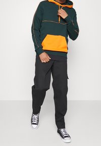 Zielona bluza z kapturem, z pomarańczową przednią kieszonką i akcentami, z kapturem na sznurki, w zestawie z luźnymi czarnymi spodniami i czarnymi sneakersami z wysoką cholewką.