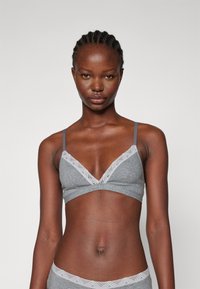 Grå bralette med justerbara axelband, V-ringning och spetsdekoration längs kanten. Mjuk tyg och sömlös design ger komfort.