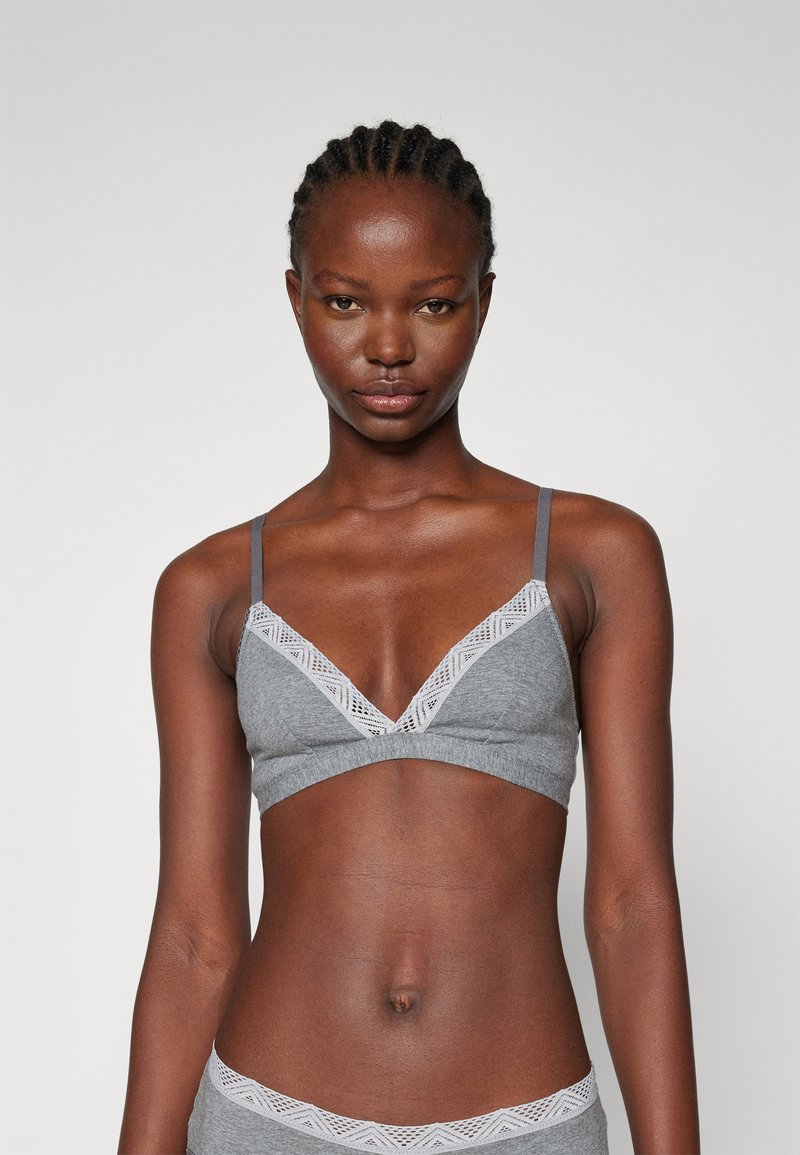 Grå bralette med justerbara axelband, V-ringning och spetsdekoration längs kanten. Mjuk tyg och sömlös design ger komfort.