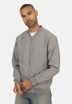 Uomo che indossa una giacca bomber grigio chiaro con cerniera sopra una maglietta bianca, mentre sistema il polsino con entrambe le mani, in piedi davanti a uno sfondo chiaro e uniforme.