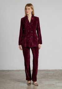 Mujer de pie vestida con un blazer de terciopelo burdeos oscuro de doble botonadura y pantalones a juego, usando tacones beige sobre un suelo gris.