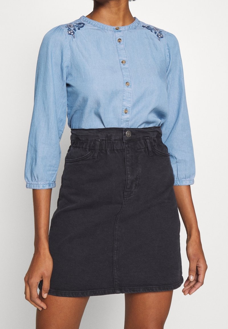 Femme portant une blouse bleu clair boutonnée avec des épaules brodées et une jupe en jean noire taille haute avec des poches devant.