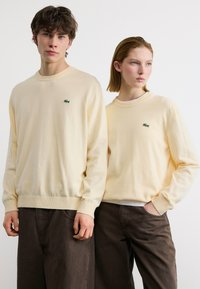 Deux jeunes adultes portant des sweat-shirts crème assortis avec un logo de crocodile vert, se tenant devant un fond blanc, tous deux regardant droit devant.