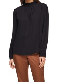 Schwarze Bluse mit langen Ärmeln, hohem Kragen und plissierten Details, aus leichtem Stoff, kombiniert mit braunen Satinhosen.
