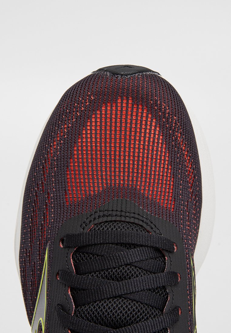 Vista superior de una zapatilla de correr de malla negra y roja con cordones negros y un acento amarillo sobre un fondo gris claro.