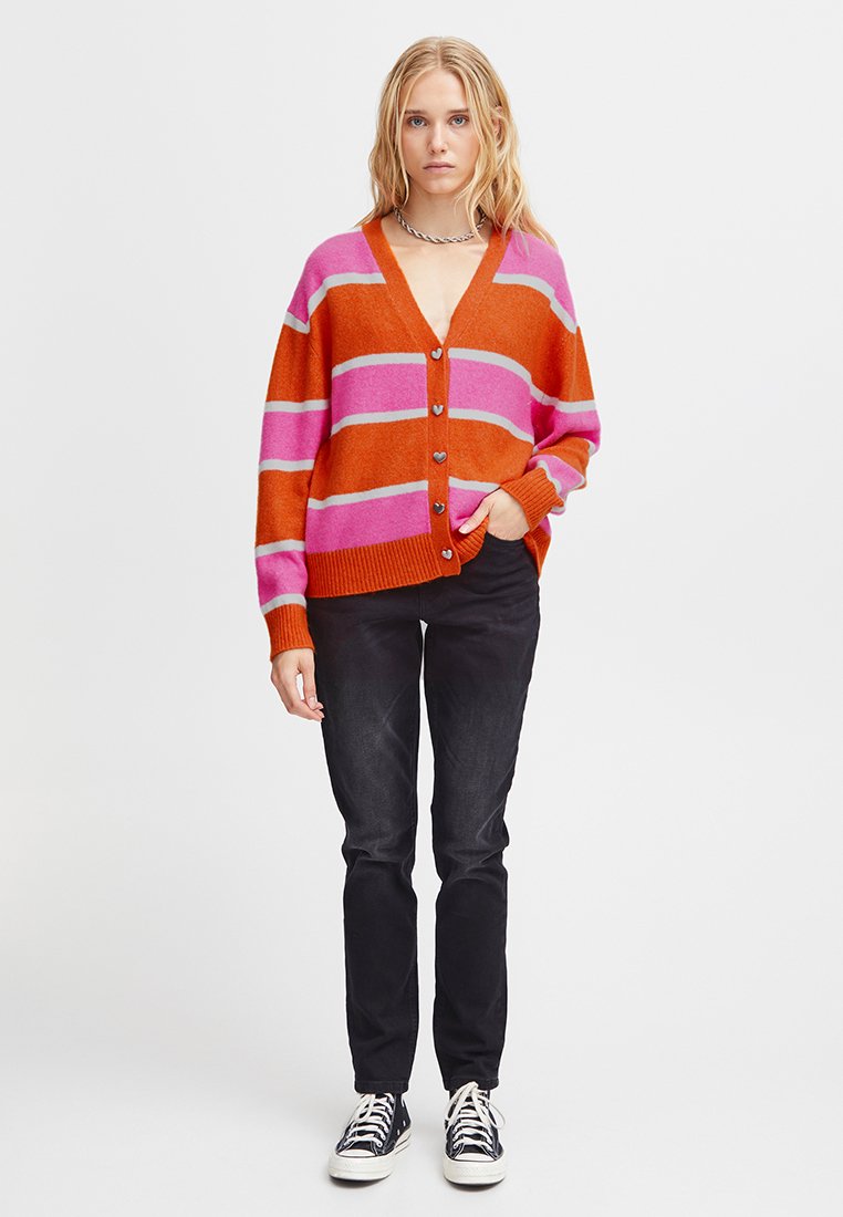 Blondynka w różowym, pomarańczowym i czerwonym paskowanym cardiganie zapinanym na guziki, czarnych dżinsach i czarnych sneakersach typu high-top, stoi z jedną ręką w kieszeni.