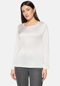 Hvid bluse med lange ærmer og firkantet udskæring, glat tekstur og sideslids ved manchetterne, kombineret med grå slim-fit bukser.