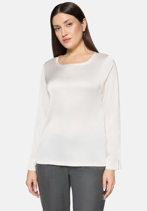 Weiße Langarmbluse mit quadratischem Ausschnitt, glatter Textur und seitlichen Schlitzen an den Manschetten, kombiniert mit grauen Slim-Fit-Hosen.