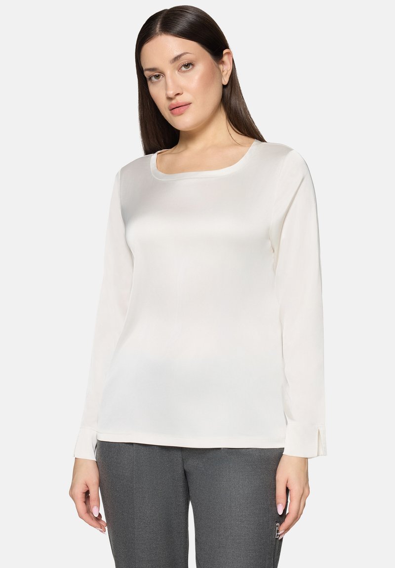 Hvid bluse med lange ærmer og firkantet udskæring, glat tekstur og sideslids ved manchetterne, kombineret med grå slim-fit bukser.