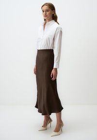 Gonna midi in satin marrone abbinata a una camicia bianca con bottoni e décolleté beige. Texture liscia e silhouette fluida.