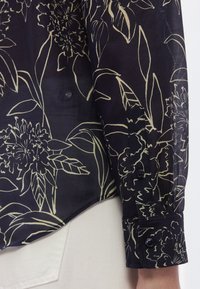 Camisa preta com estampa floral e textura transparente, com mangas compridas, punhos com botões e linha inferior curva, em contraste com calças de cor clara.